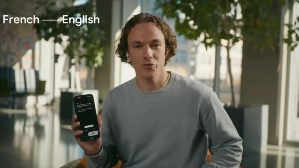 A man in a gray sweater holds up a phone that's doing live translation of French to English language. He's wearing earbuds.