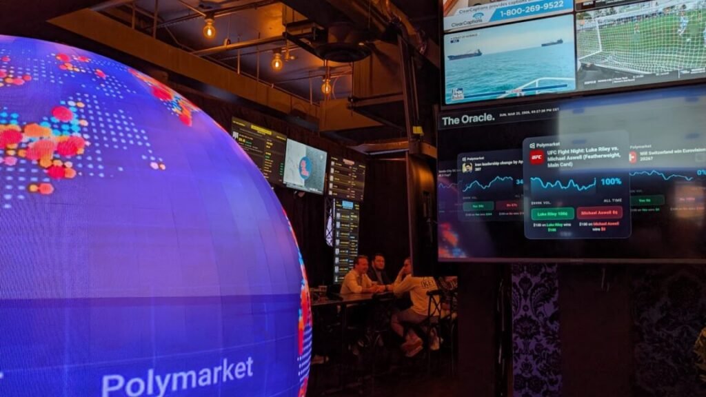 Pints meet prop bets: Polymarket’s “Scenario Room” pop-up bar in DC Image of group of people sitting between a blue globe and a TV screen showing a mixture of sports and current events.