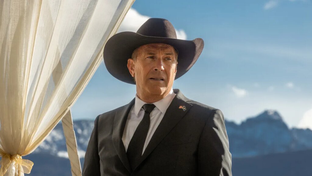 John Dutton, played by Kevin Coster, stands in his cowboy hat and suit with Montana mountains in the background.