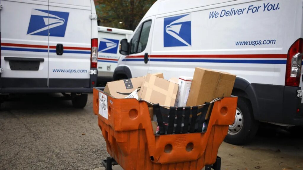 An orange cart filled with packages in front of three postal service trucks.