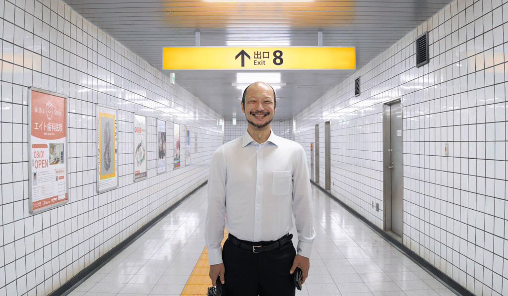 Yamato Kochi as The Walking Man, standing wearing a white shirt, black trousers and grinning at the viewer. He is standing in a subway tunnel.