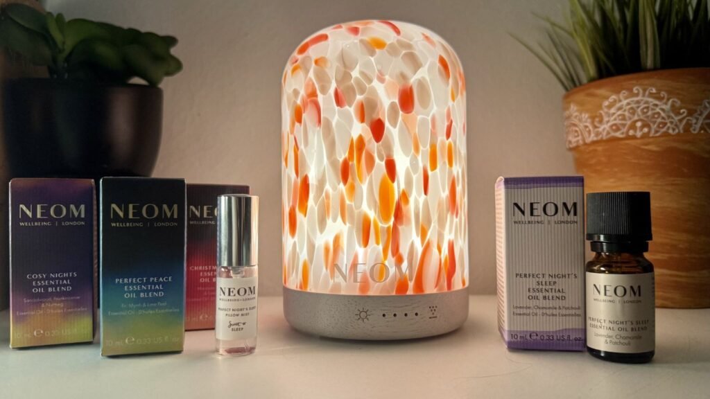 NEOM Clementine Wellbeing Pod (pink, orange and white speckle diffuser) next to bottles and boxes of NEOM essential oil blends including the Perfect Night's Sleep blend (purple and white box).