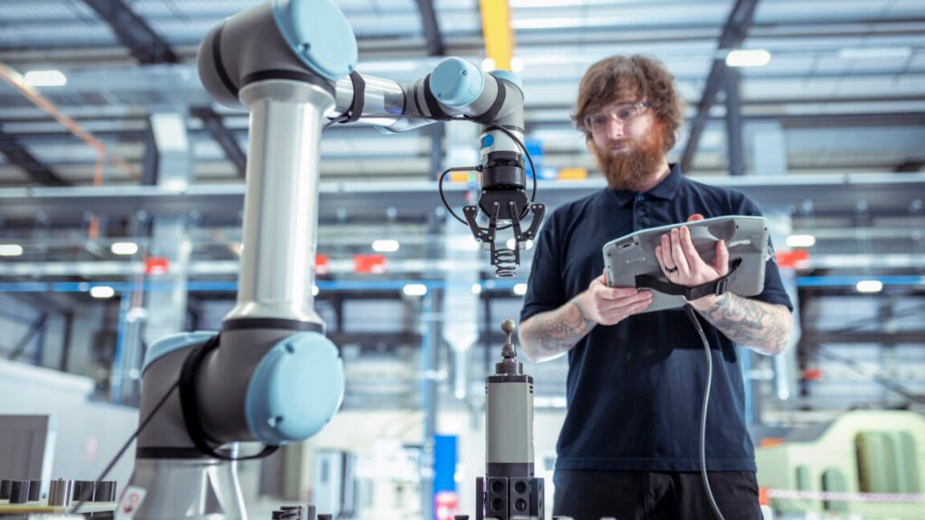 Image of a man with a computer tablet standing in front of an industrial robotic arm.