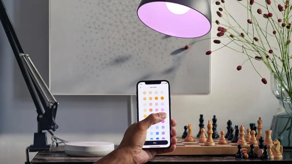 A user controls a smart lamp with their phone. A Samsung hub, which is required, is on a table in the background.
