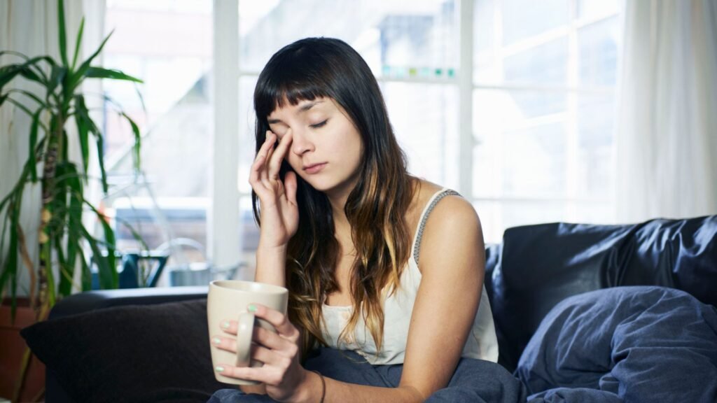 Sleep specialists clarify how this morning behavior is draining your vitality A woman sis on her couch looking tired, with her eyes closed. She has a cup of coffee in her hand.