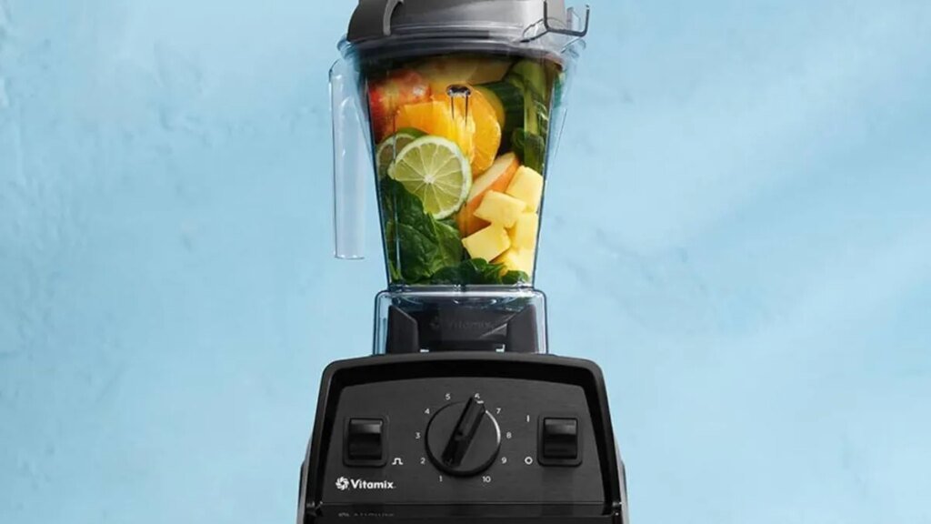 The Vitamix E310 Explorian blender filled with fruit and greens. The background is light blue.